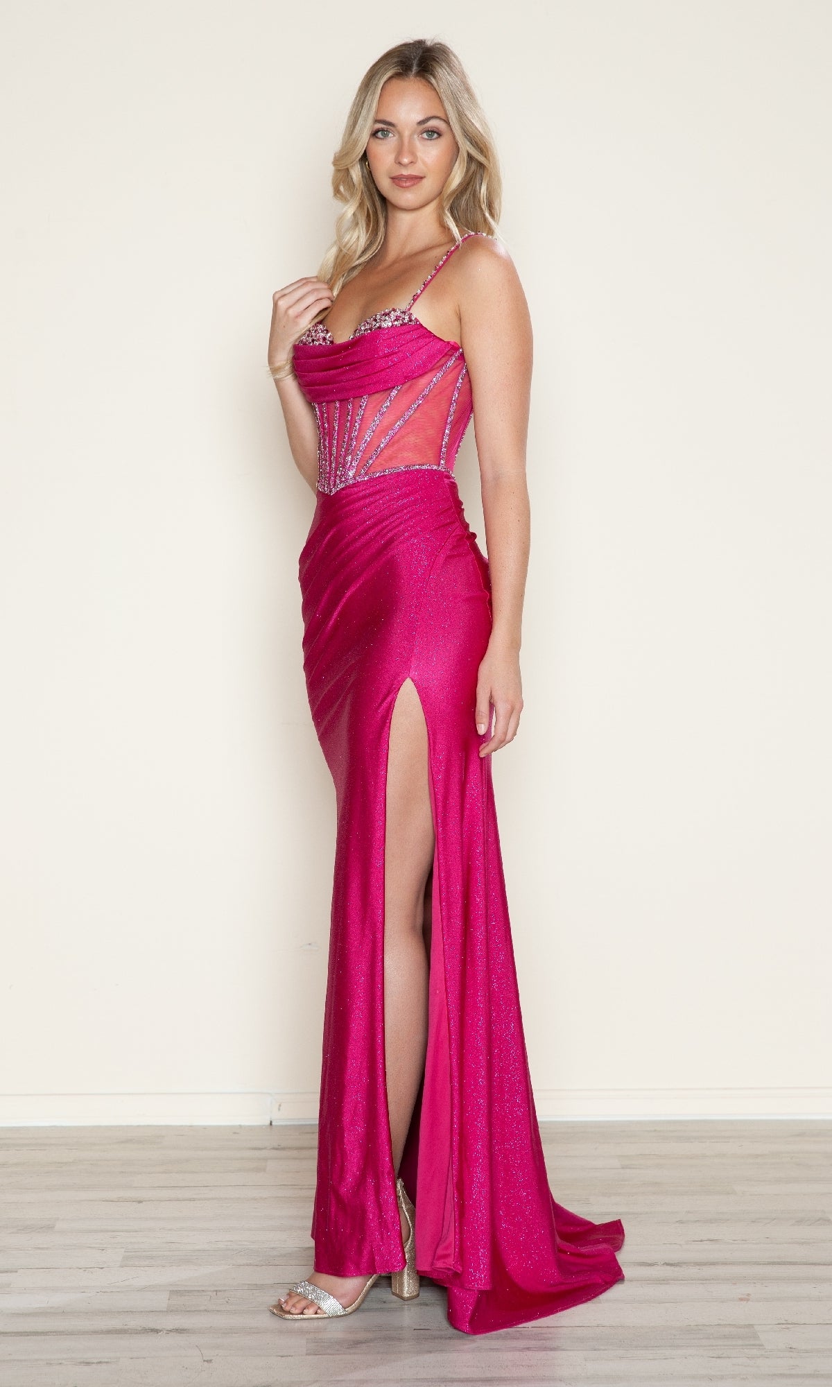 A woman stands indoors wearing the Sheer Beaded-Corset Long Prom Dress: Poly USA 9750, featuring a bright pink hue, beaded sheer bodice, floor-length skirt with a thigh-high slit, and strappy heels.